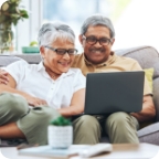 A senior couple sit together on their couch and use a laptop to compare insurance policies. 